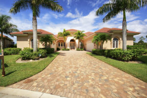 Luxurious single-story home with terracotta roof, manicured garden, and palm trees. The stone driveway leads to a grand entrance under a blue sky.
