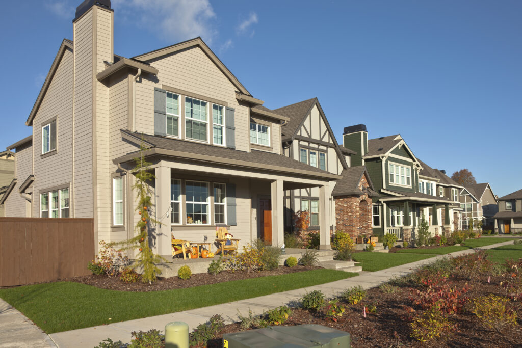 Row of suburban houses under a clear blue sky, featuring manicured lawns and autumn decorations on porches, evoking warmth and community.