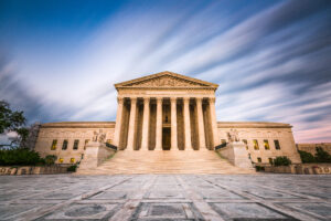 Grand, neoclassical building with tall columns and ornate pediment under a dynamic sky. The wide plaza enhances its imposing presence.